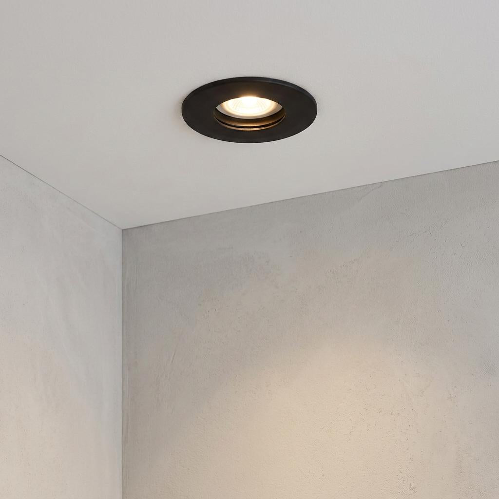 Black circular recessed downlight with warm light installed in a white ceiling above grey walls.
