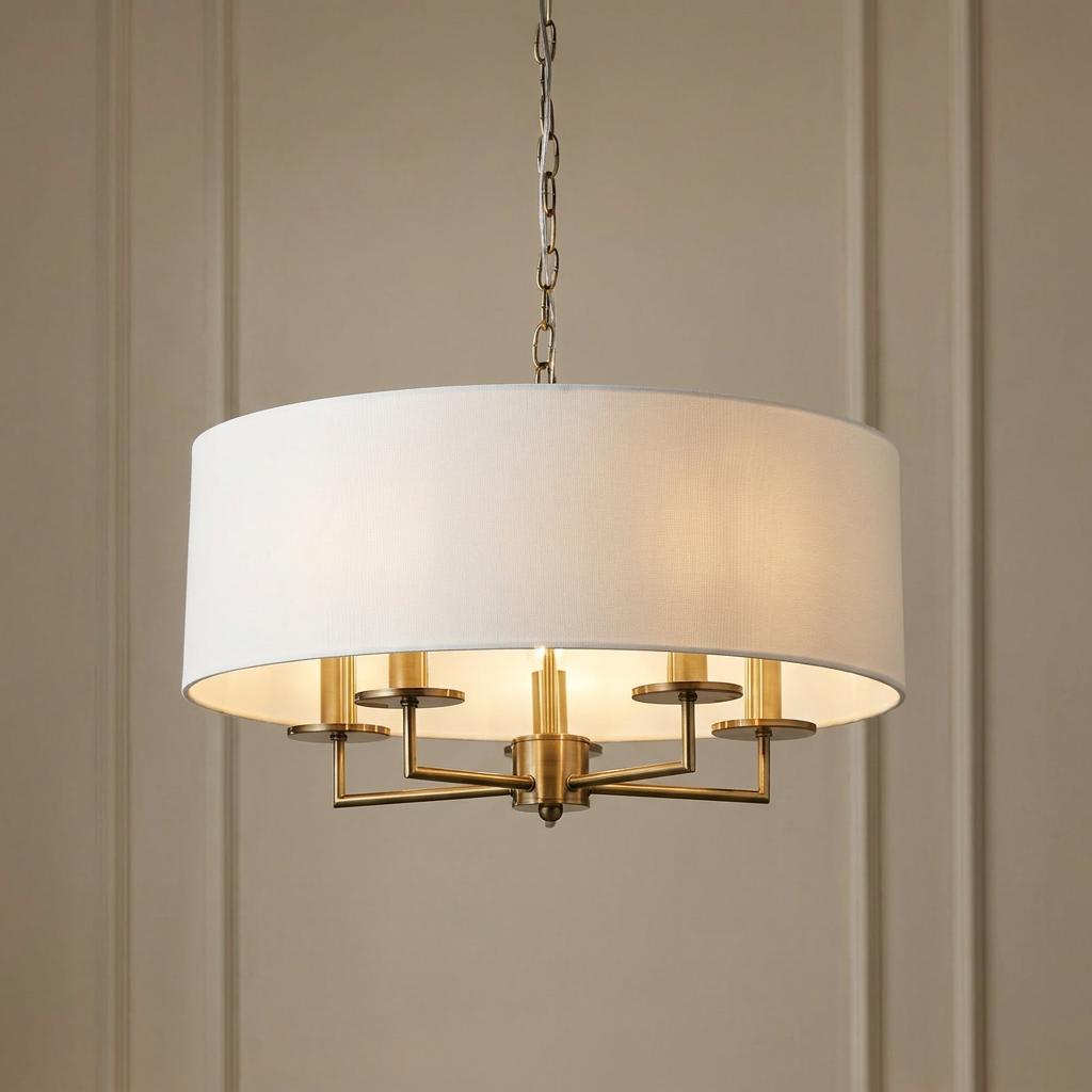 Five-light antique brass pendant chandelier featuring a white linen drum shade, lit against a paneled wall.