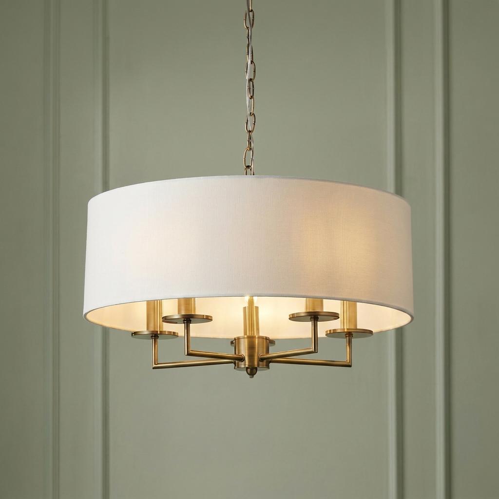 Five-light brass pendant chandelier with a white linen drum shade, set against a green paneled wall.