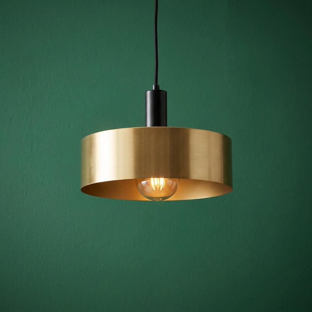 Brushed gold cylindrical pendant light with a black fitting and glowing bulb against a dark green textured wall.