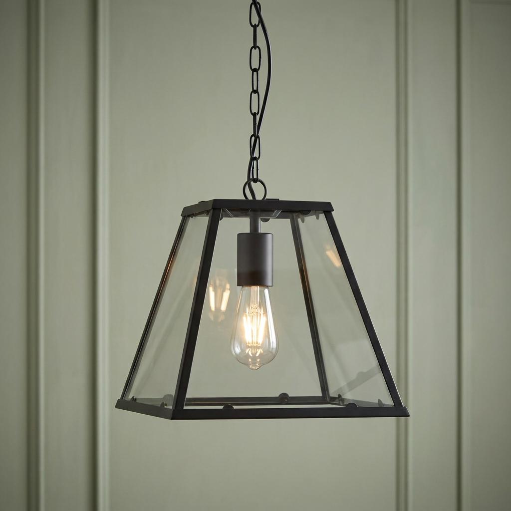 Black lantern pendant light with clear glass panels and an Edison bulb hanging against a green paneled wall.