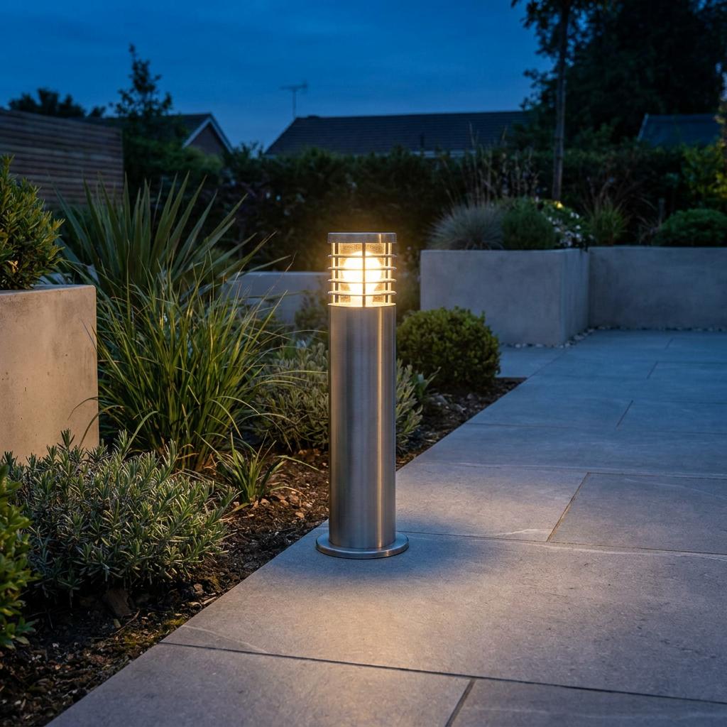 Illuminated stainless steel bollard light with a louvred top on a modern stone garden patio at dusk.