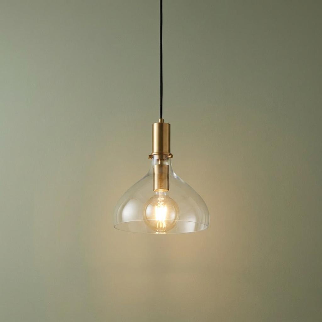 Brass pendant light with a clear glass bell-shaped shade and glowing bulb against a sage green wall.