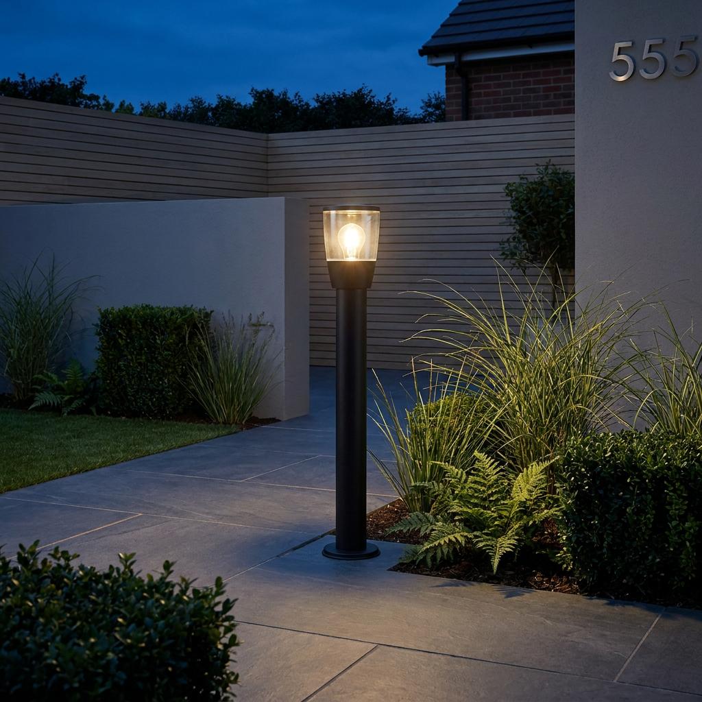 Black outdoor bollard light with a glowing glass top illuminating a modern stone garden path at dusk.