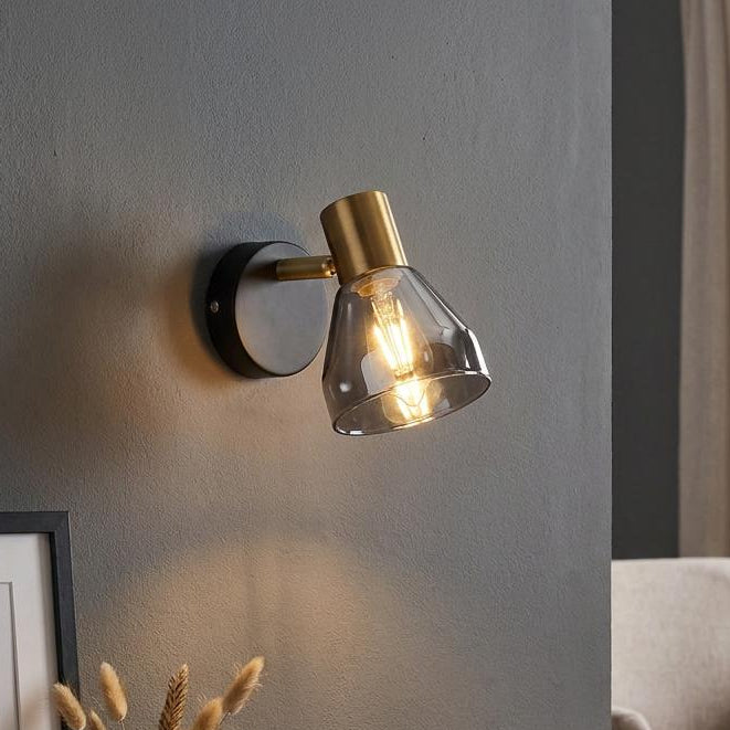 Modern black and brass wall-mounted spotlight with a smoked glass shade illuminating a grey wall.