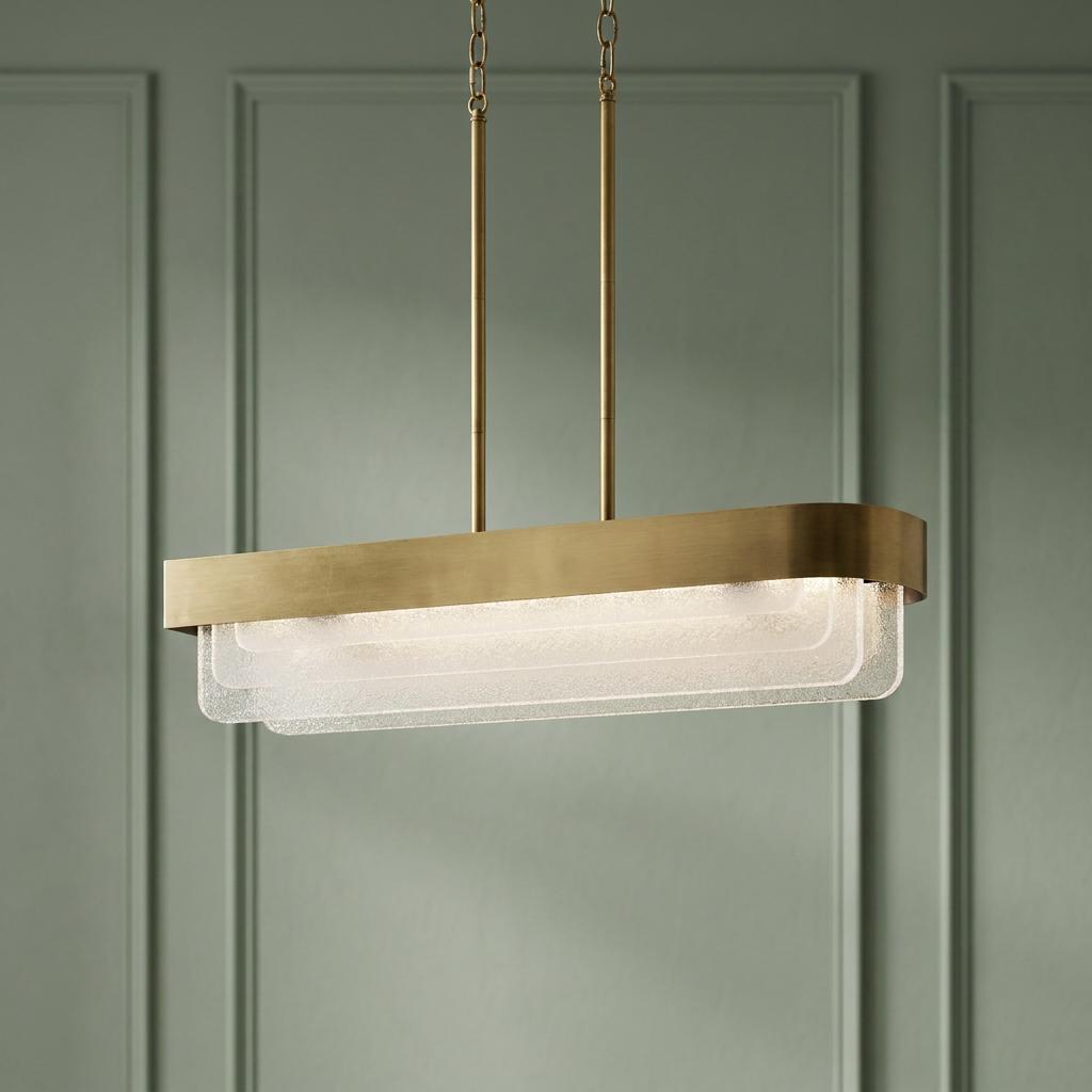 Rectangular brass LED chandelier with tiered textured glass panels hanging against a sage green paneled wall.