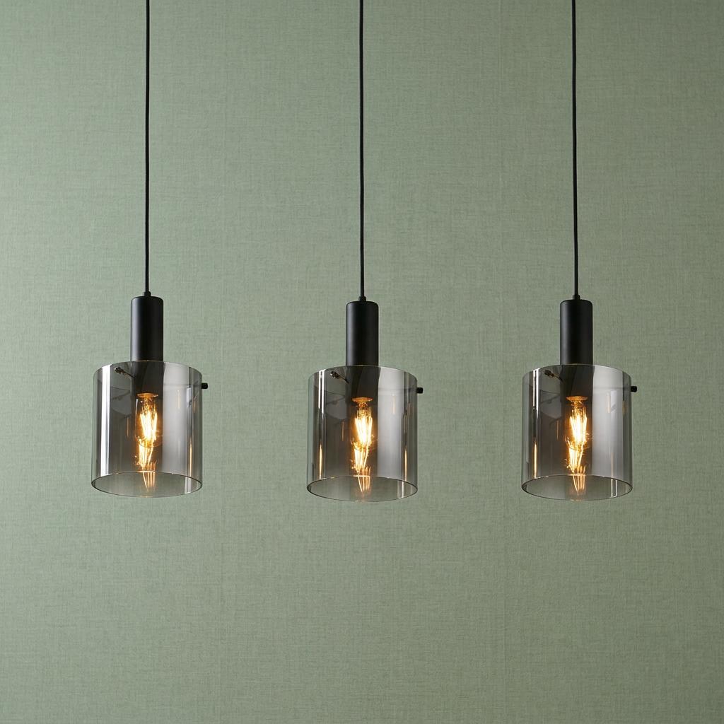 Three matte black pendant lights with smoked ombre glass cylinder shades against a green textured wall.