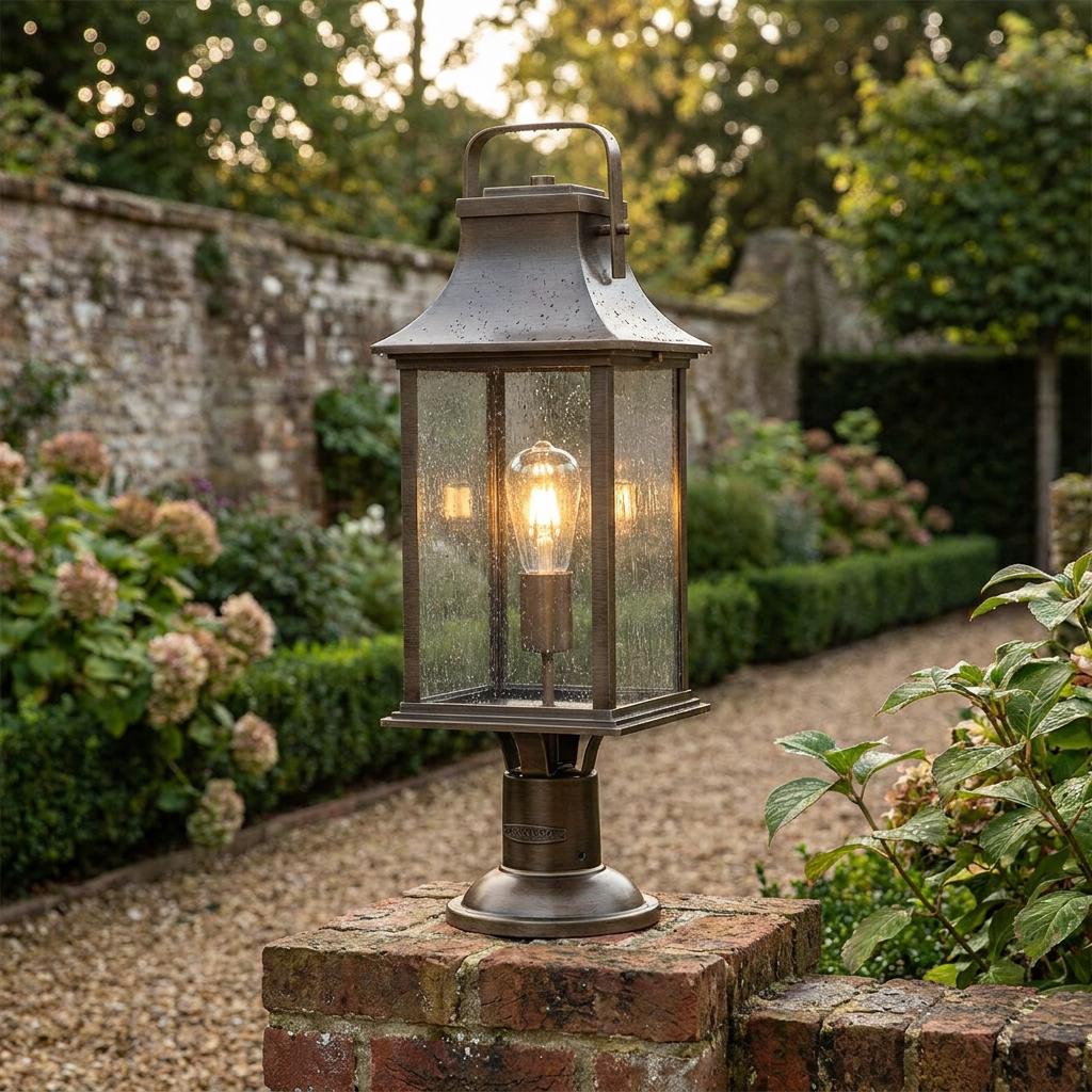 Traditional bronze lantern with seeded glass and a glowing bulb, mounted on a brick pillar in a scenic garden.