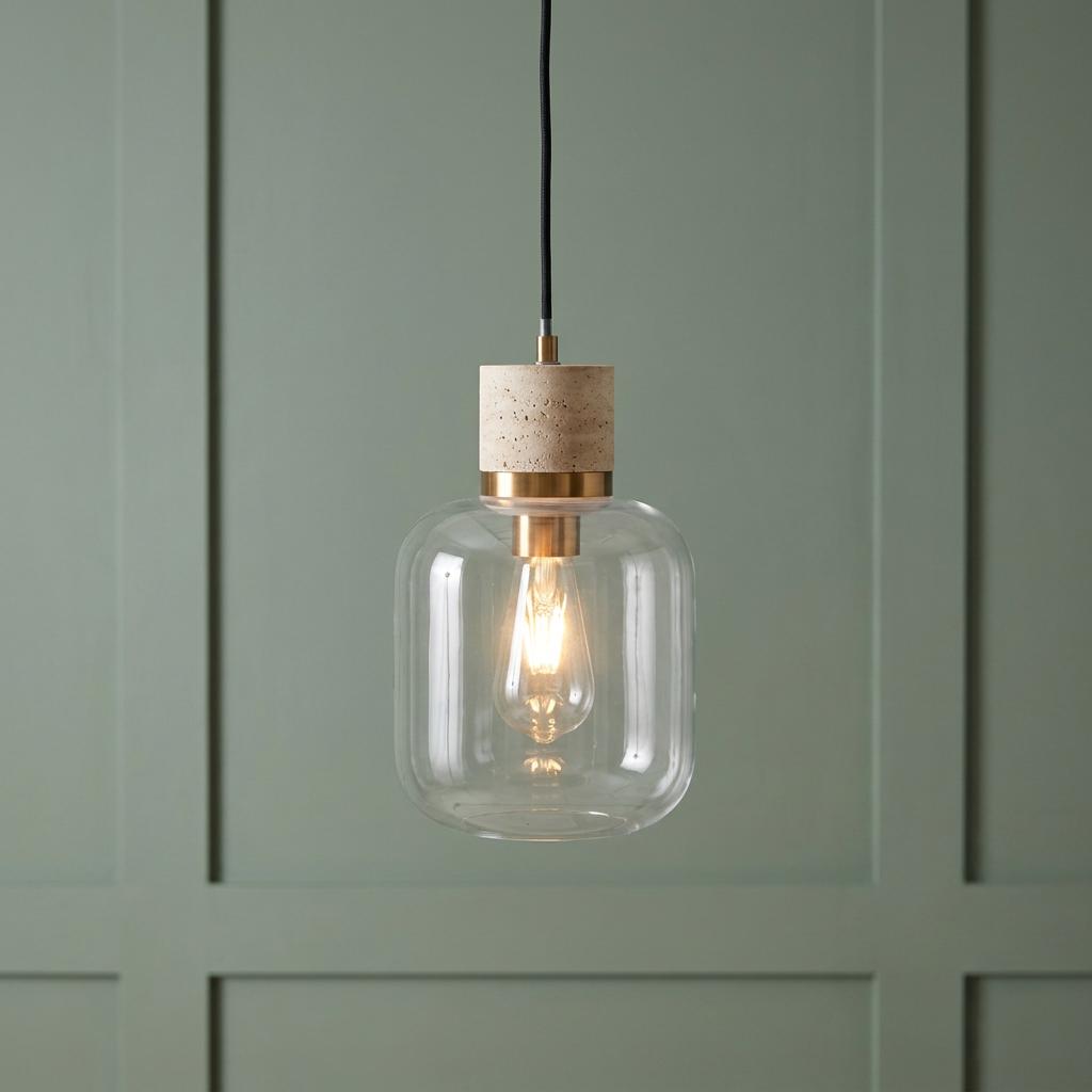A modern pendant light with a travertine stone top, brass accent, and clear glass shade against a sage green wall.