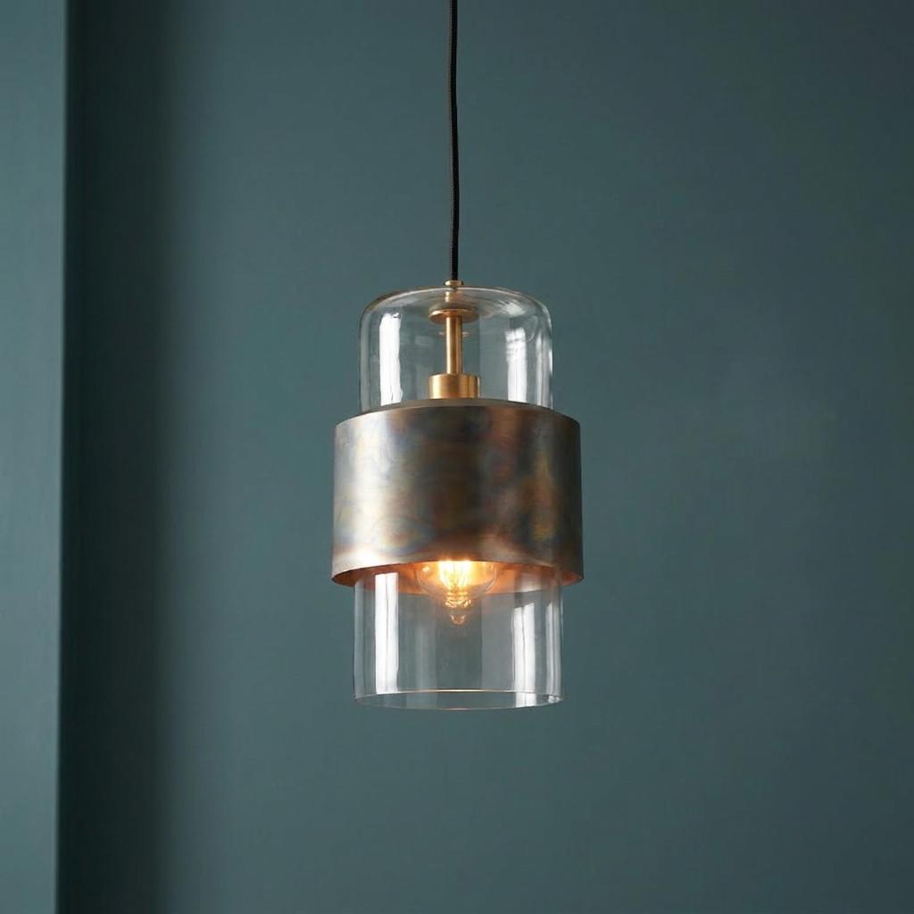 Modern brass patina pendant with a clear glass shade and glowing bulb against a dark teal wall.
