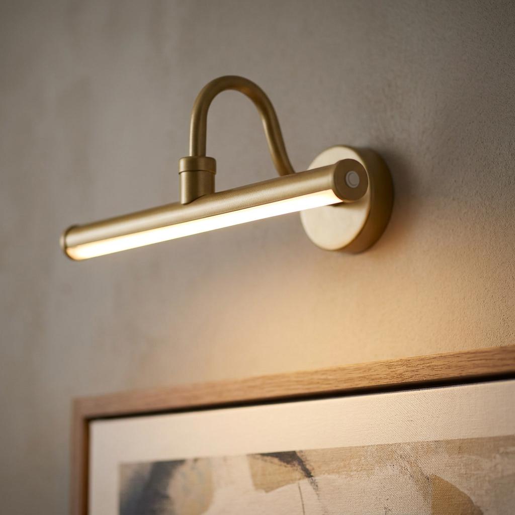 A sleek gold metal picture light mounted on a wall, casting a warm glow over a framed painting.