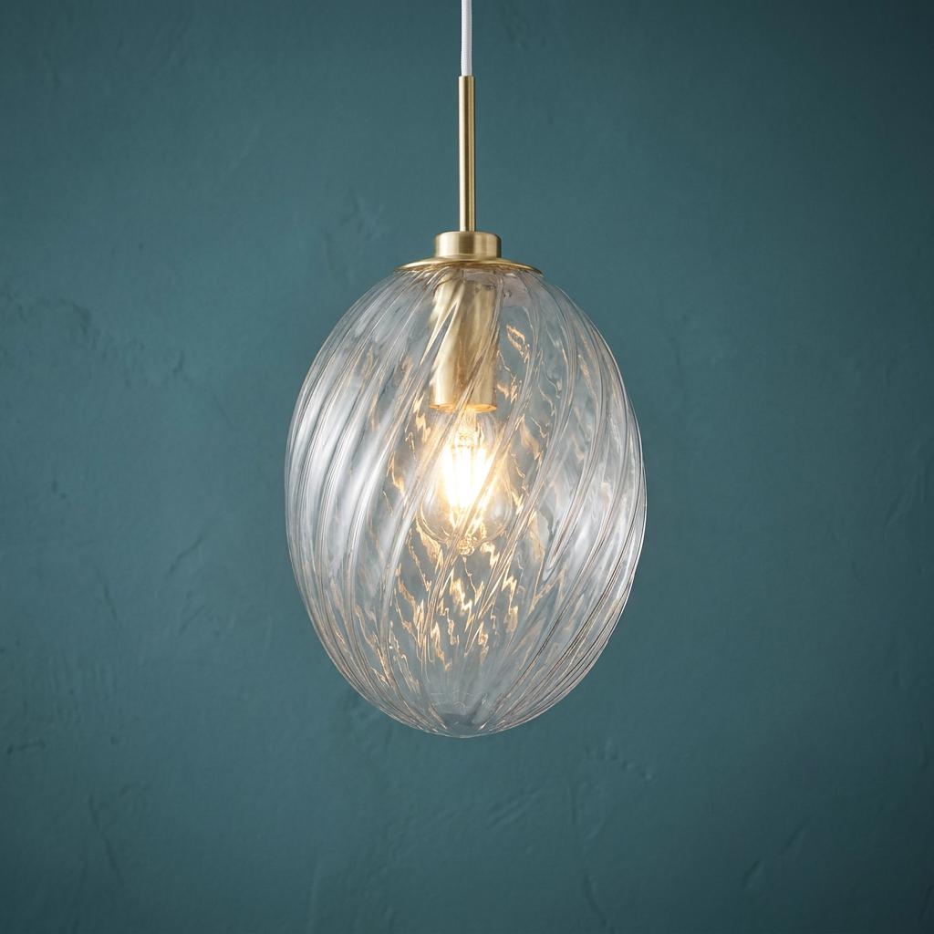 Swirled clear glass oval pendant light with satin brass hardware glowing against a textured teal wall.