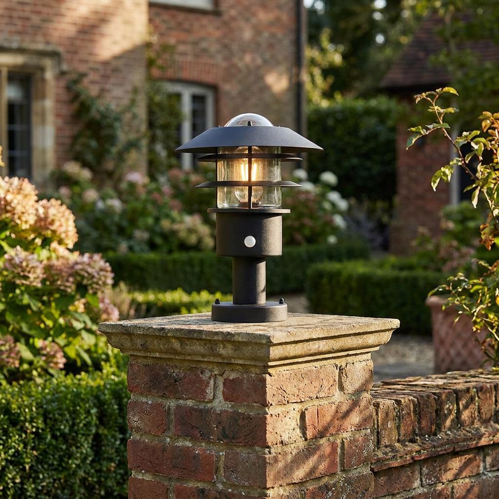 Black contemporary tiered outdoor pedestal light with PIR sensor, lit on a brick pillar in a garden.