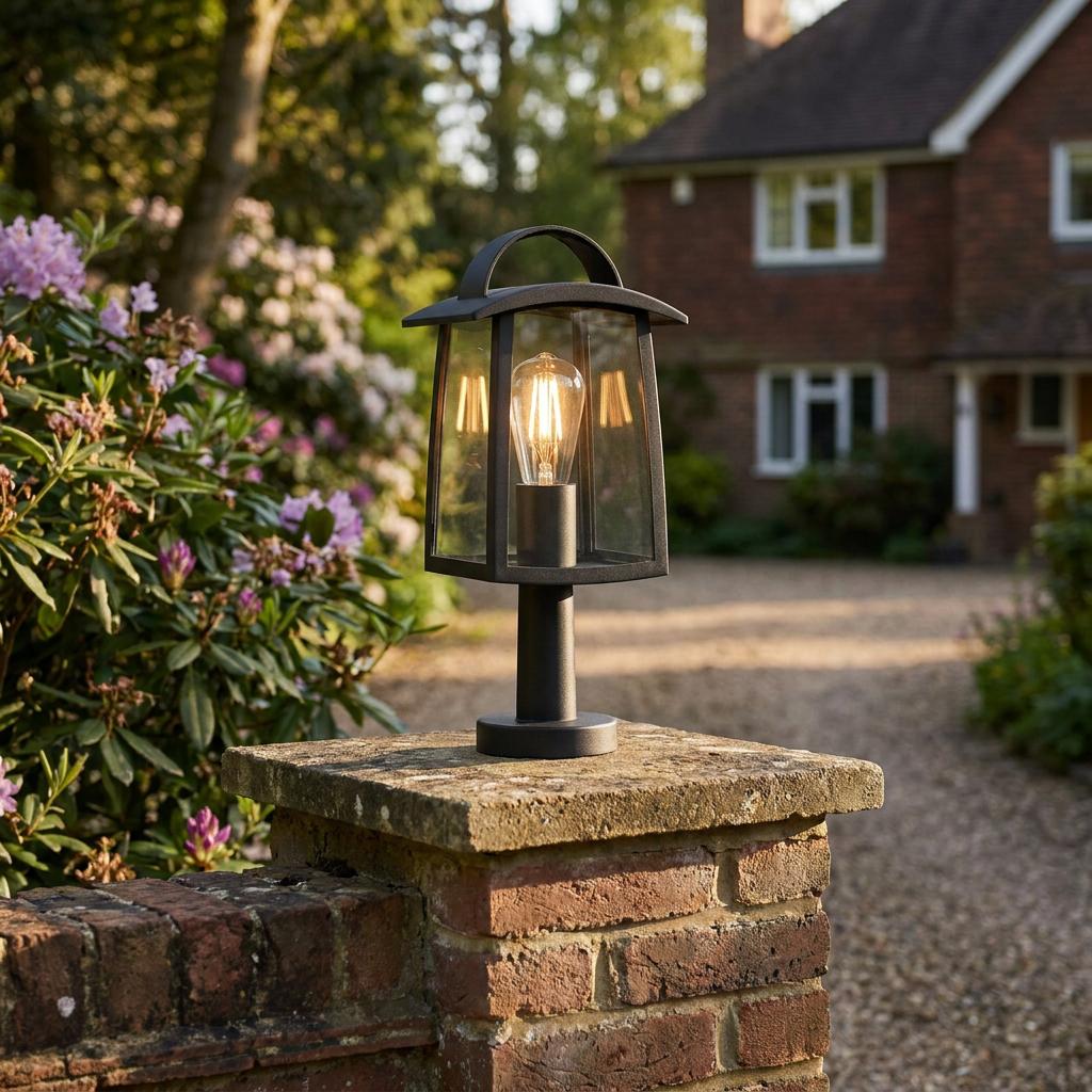 A black lantern outdoor post light with a glowing filament bulb mounted on a brick pillar in a garden.