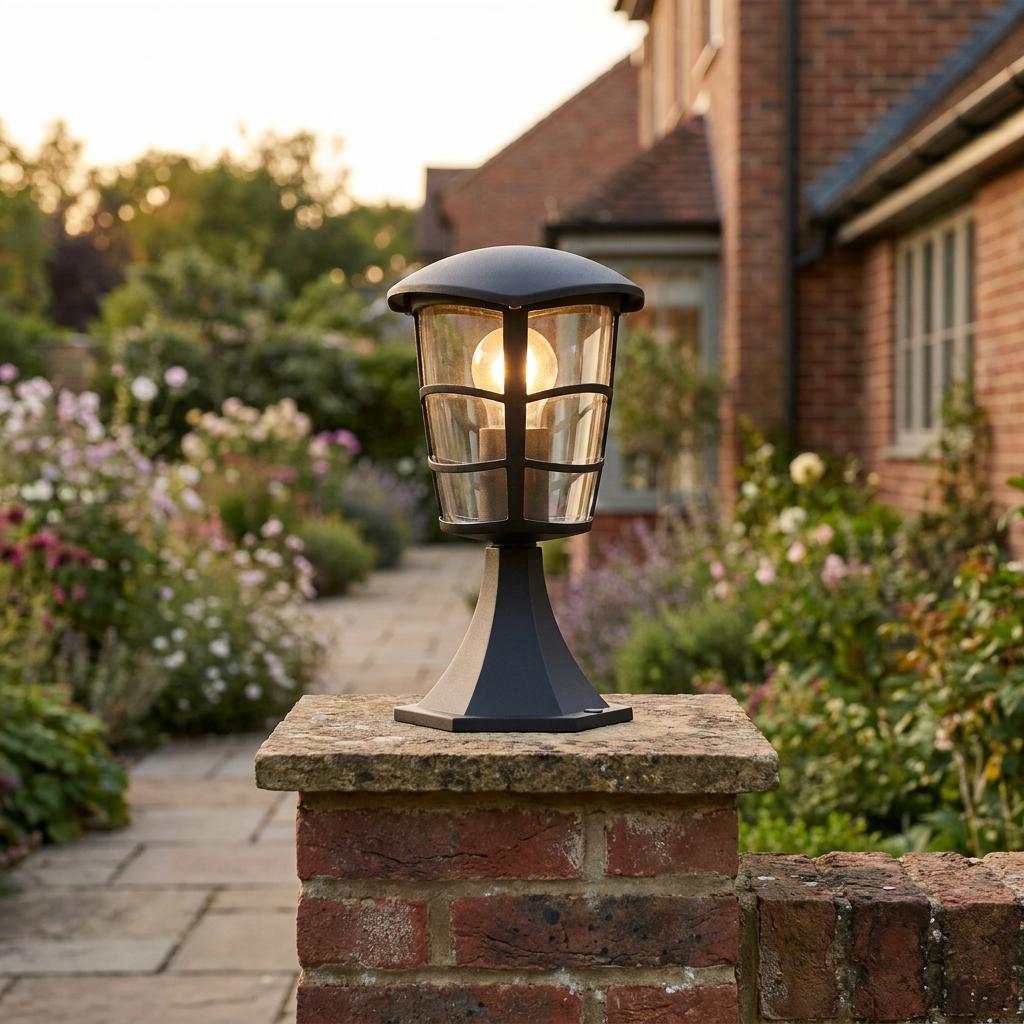 A black traditional outdoor pedestal light lit on a brick pillar in a lush garden setting at dusk.