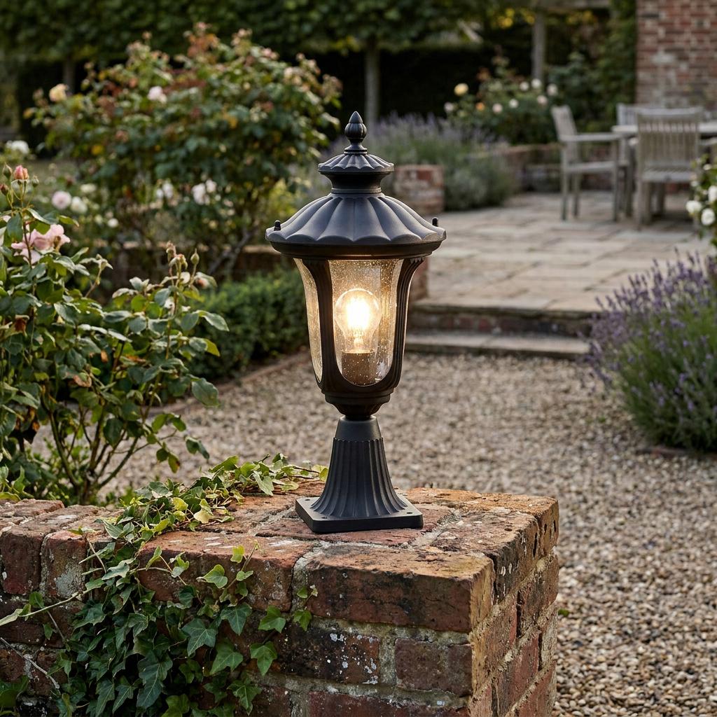 Black traditional outdoor pedestal lantern lit on a brick garden wall with a gravel path in the background.