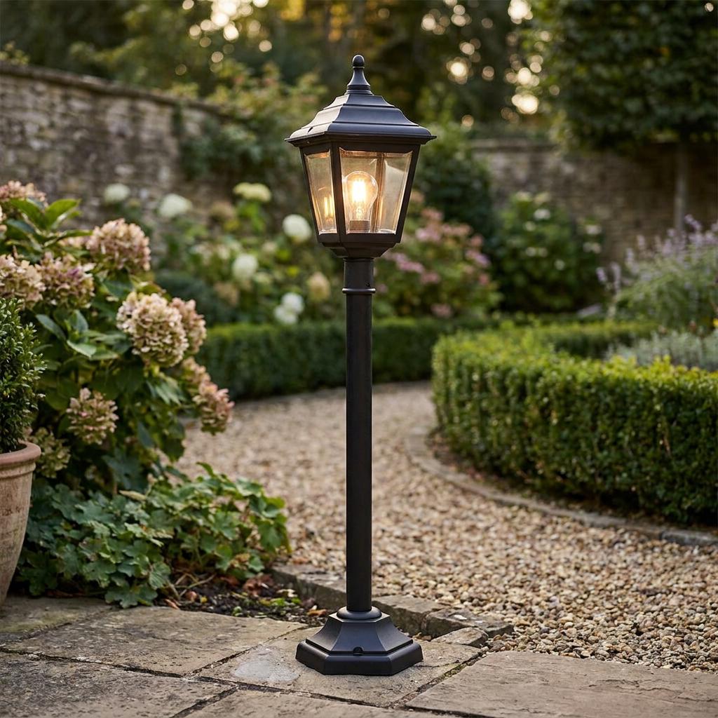Traditional black garden lamp post glowing on a gravel path surrounded by lush greenery and flowers.