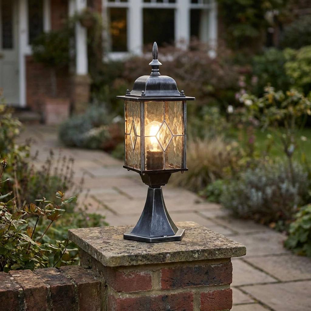 Lit lantern-style outdoor pedestal light in weathered pewter mounted on a brick pillar in a garden.