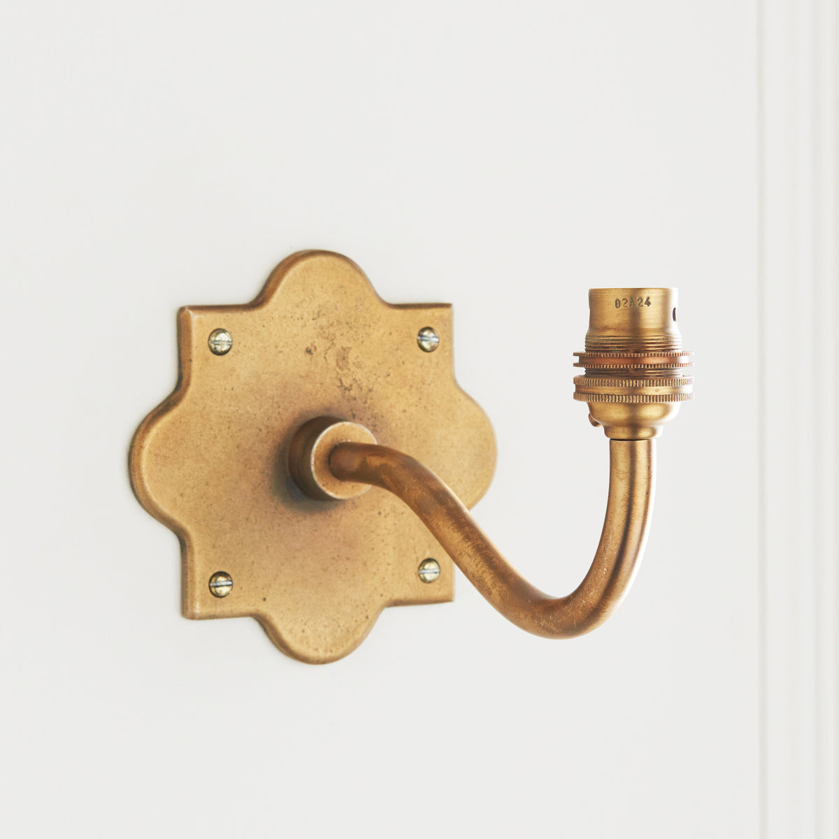 Front-angled view of the Anvil Curve wall light, its aged brass backplate screwed to white panelling