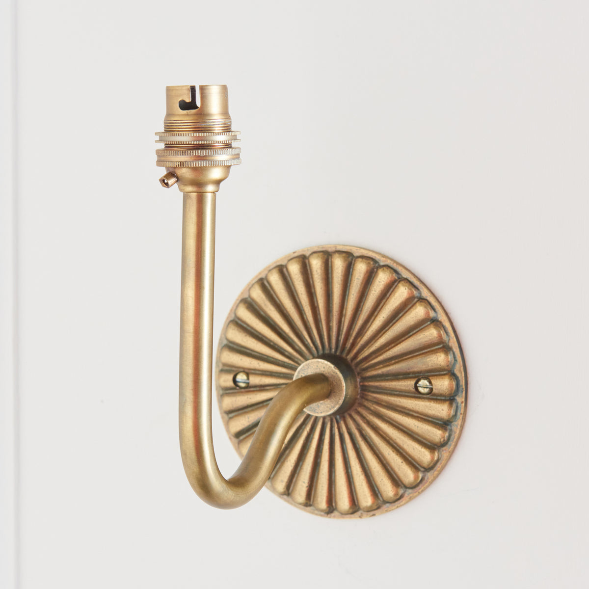 Dahlia Swan antique brass wall light on white panelling, curved arm lifting a bare brass lampholder