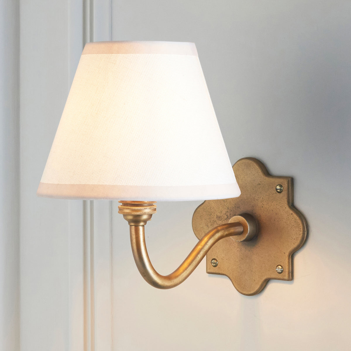 Anvil Curve brass wall light with white tapered fabric shade lit against white panelling