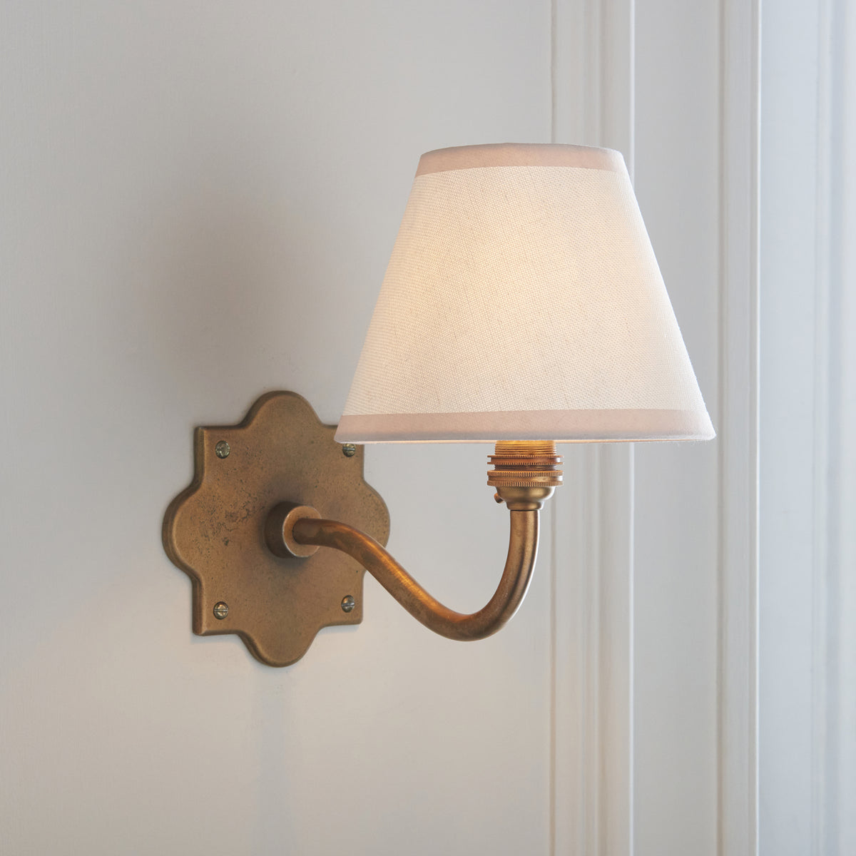 Angled view of the shaded Anvil Curve wall light, cream fabric shade above a curved brass arm
