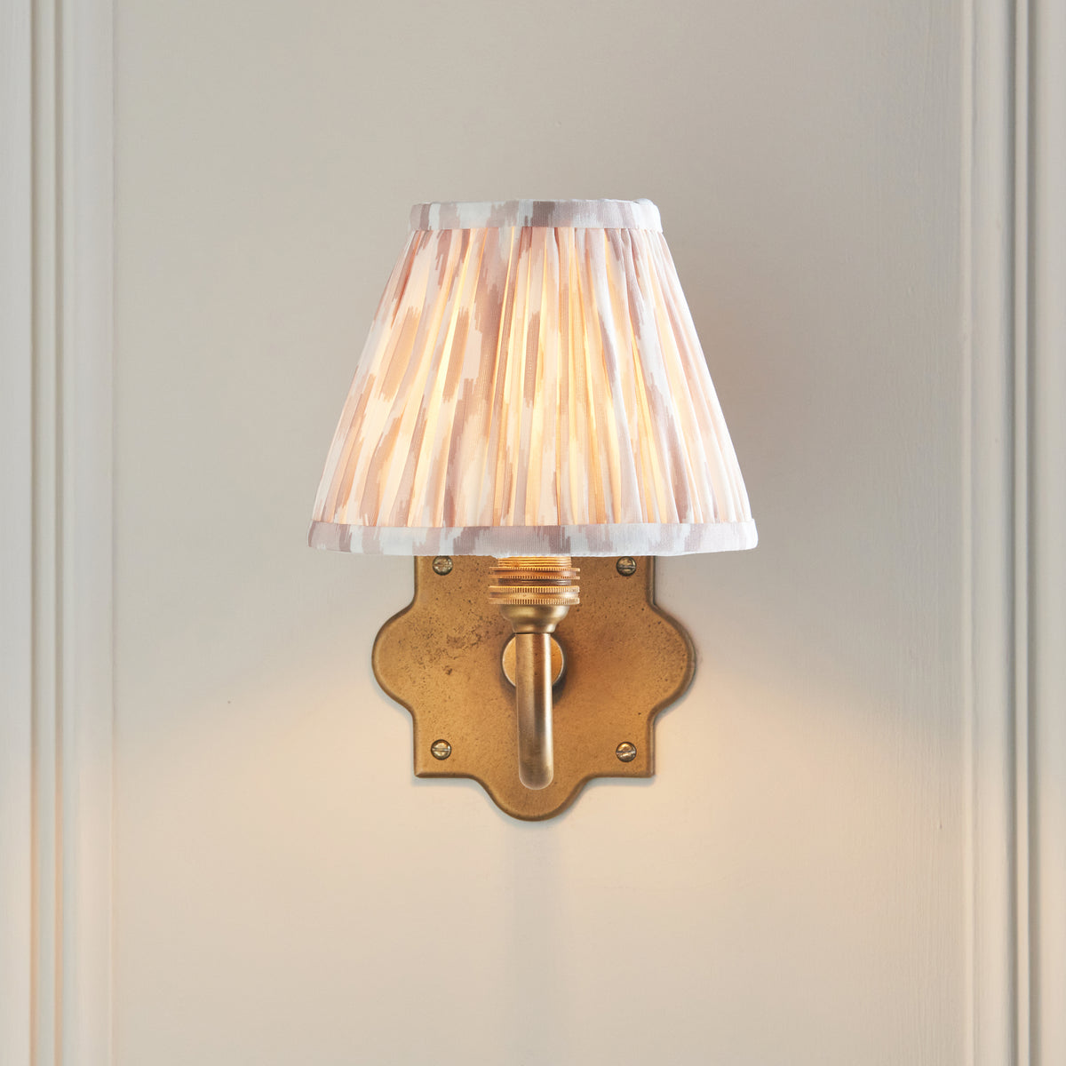 Anvil Curve wall light seen straight on, quatrefoil brass backplate below a lit pleated ikat shade