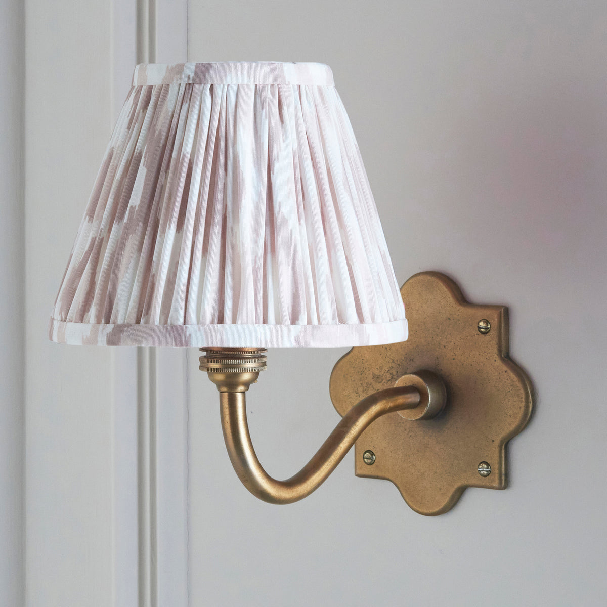 Lit Anvil Curve wall light in close view, softly glowing pleated ikat shade above aged brass arm
