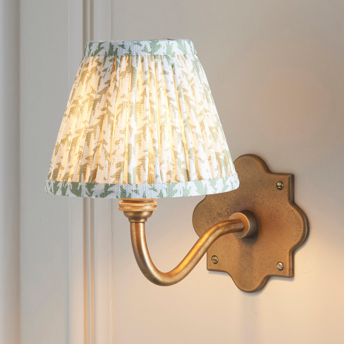 Anvil Curve brass wall light lit on cream panelling, green leaf-patterned pleated shade glowing