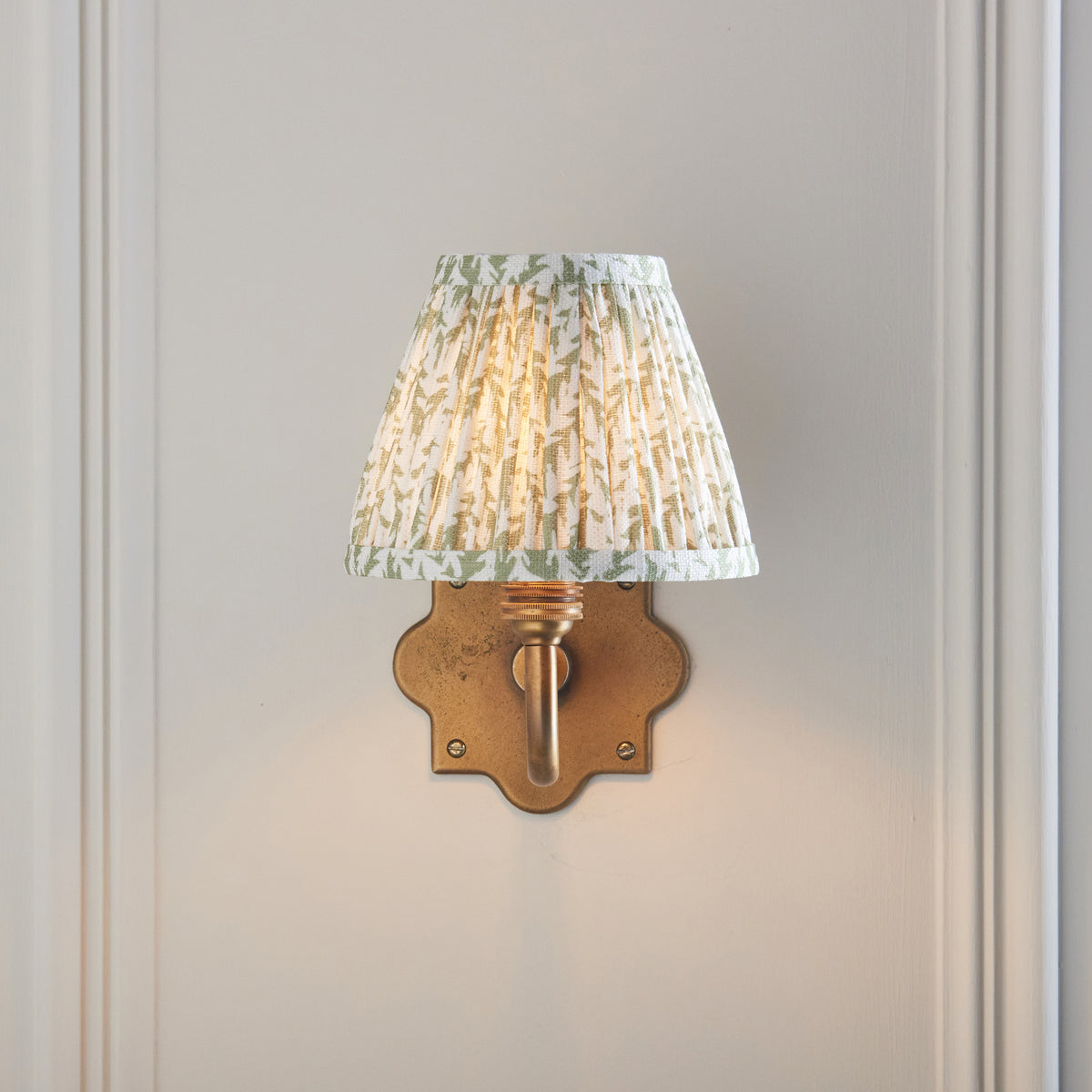 Front view of the Anvil Curve wall light, quatrefoil brass plate under a lit green leaf-print shade