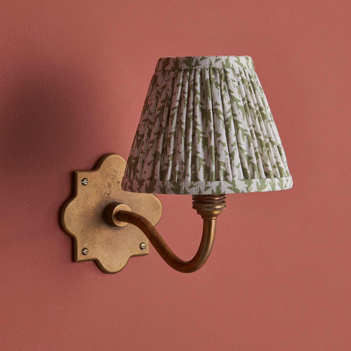 Anvil Curve brass wall light mounted on a deep terracotta wall with leafy green pleated shade lit
