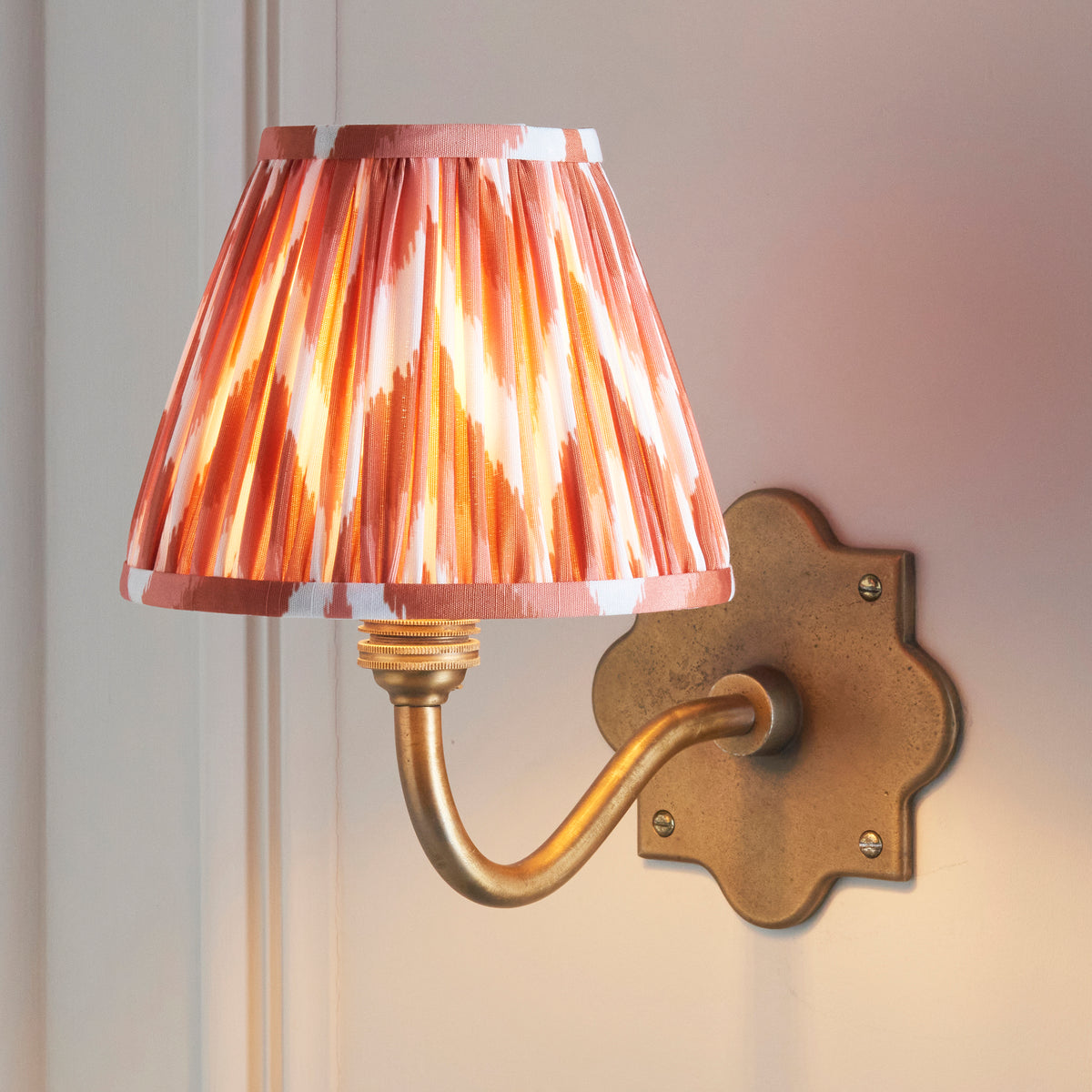 Anvil Curve brass wall light lit on panelling, coral zigzag ikat pleated shade casting a warm glow