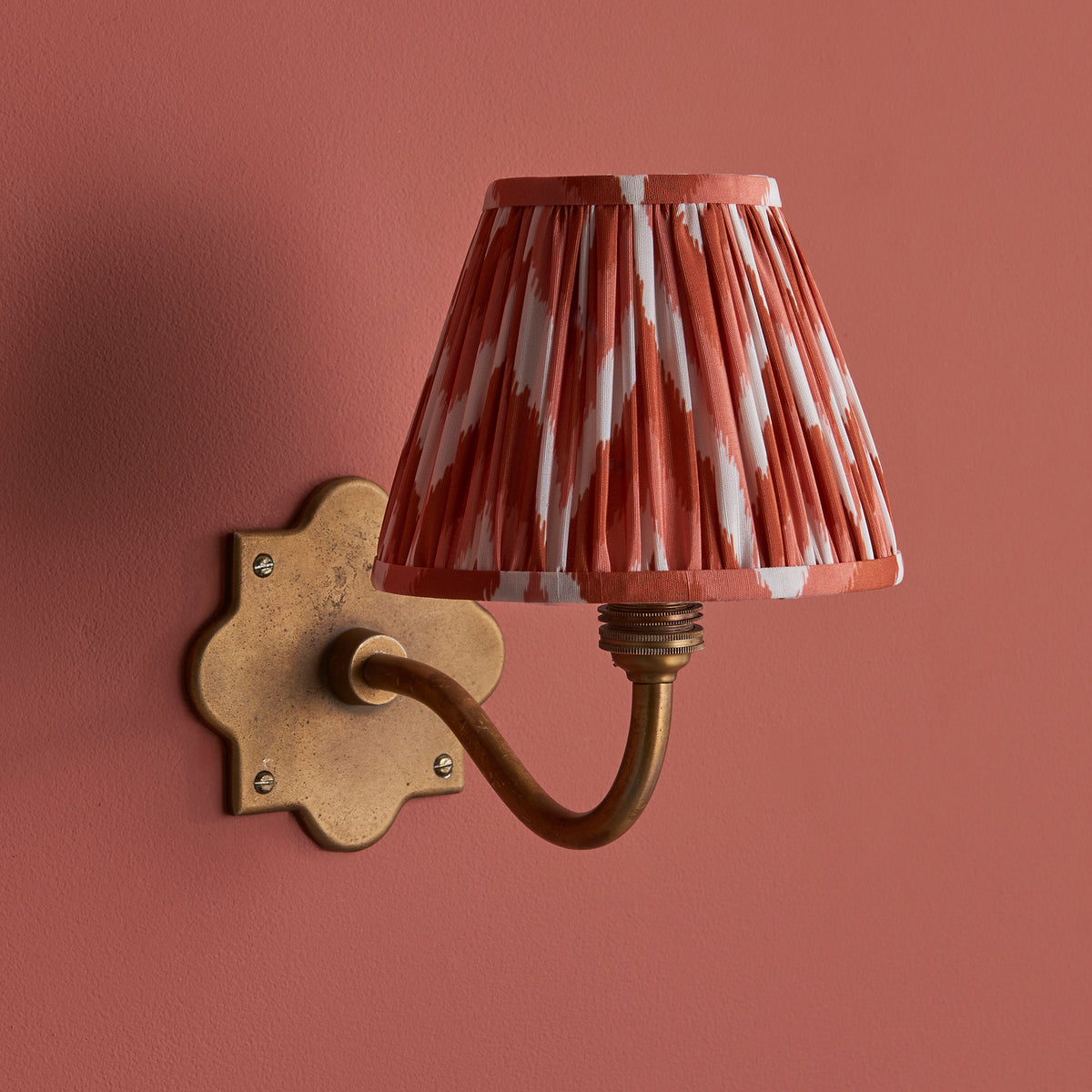 Anvil Curve brass wall light against deep terracotta paint, coral zigzag ikat pleated shade lit