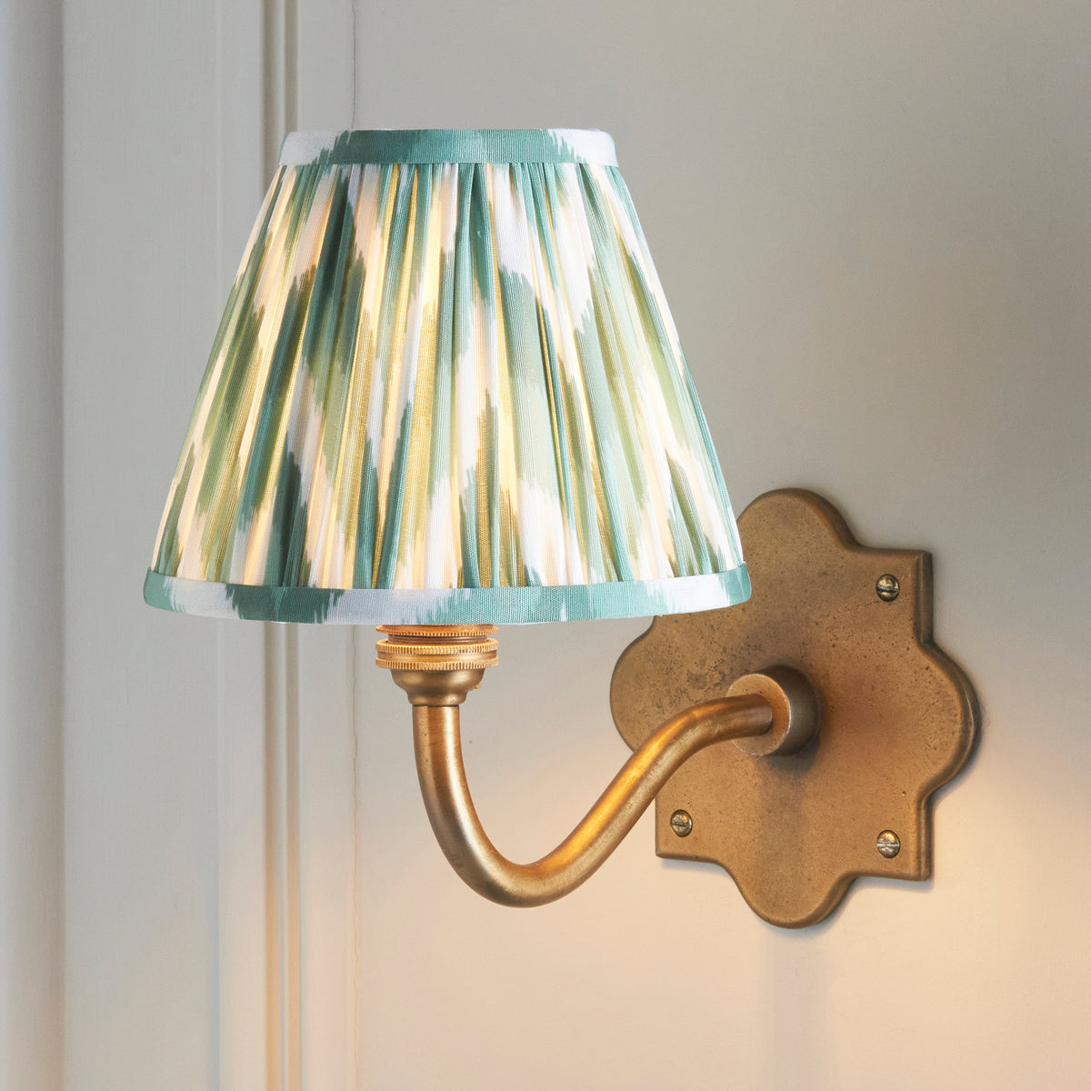 Anvil Curve brass wall light lit on cream panelling, soft green zigzag ikat pleated shade