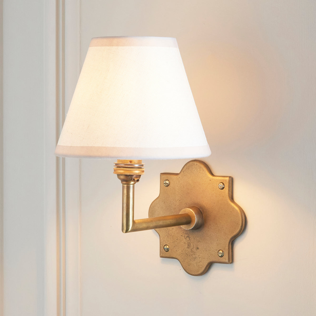 Anvil Angle brass wall light lit on cream panelling, ivory shade casting a soft glow