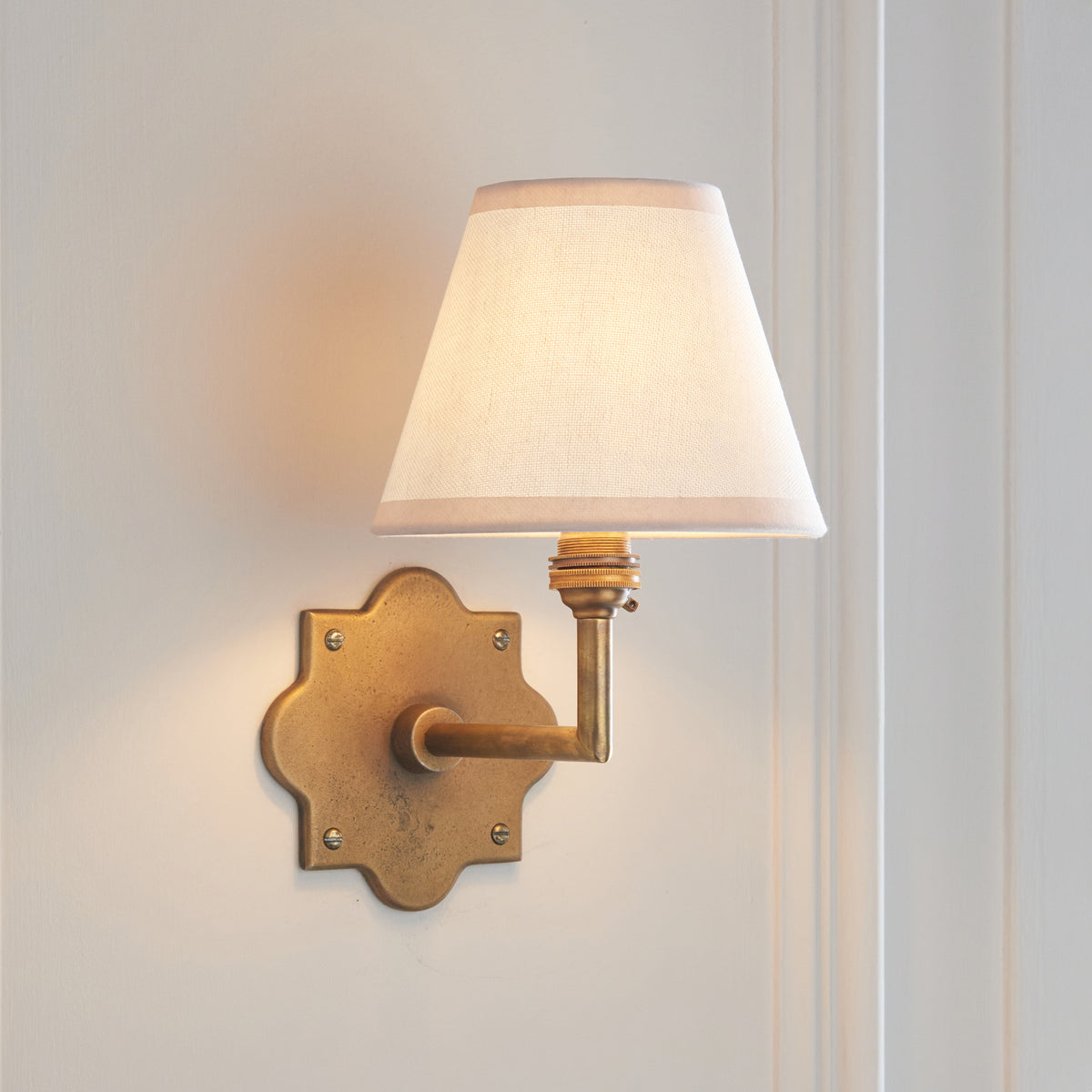 Anvil Angle brass wall light angled on panelling, warm light through the plain ivory shade