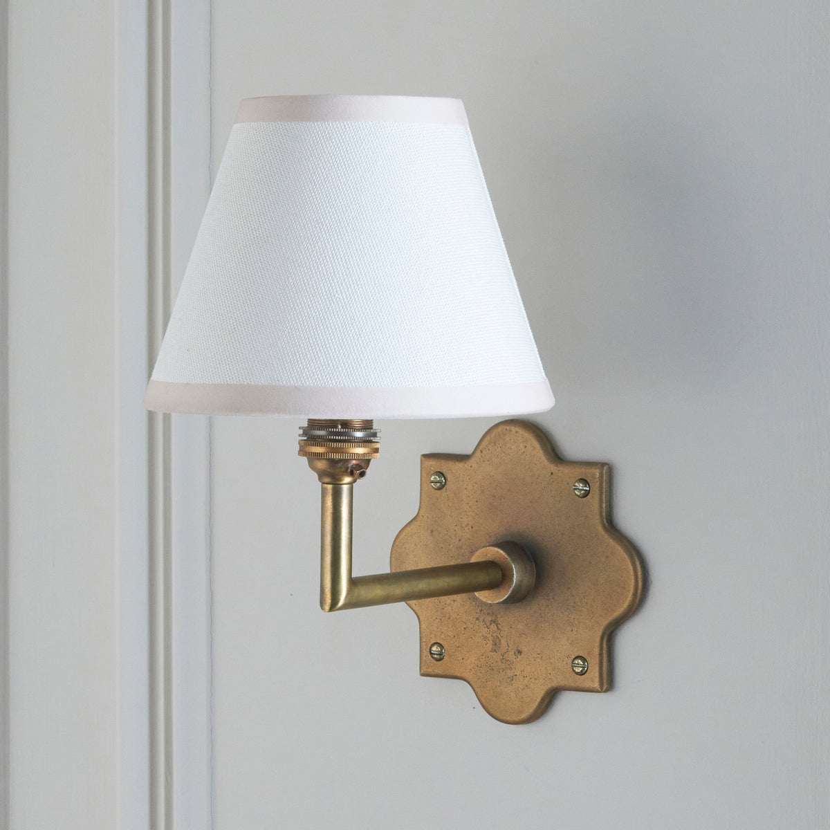 Anvil Angle wall light on grey-white panelling, crisp white tapered shade and aged brass arm