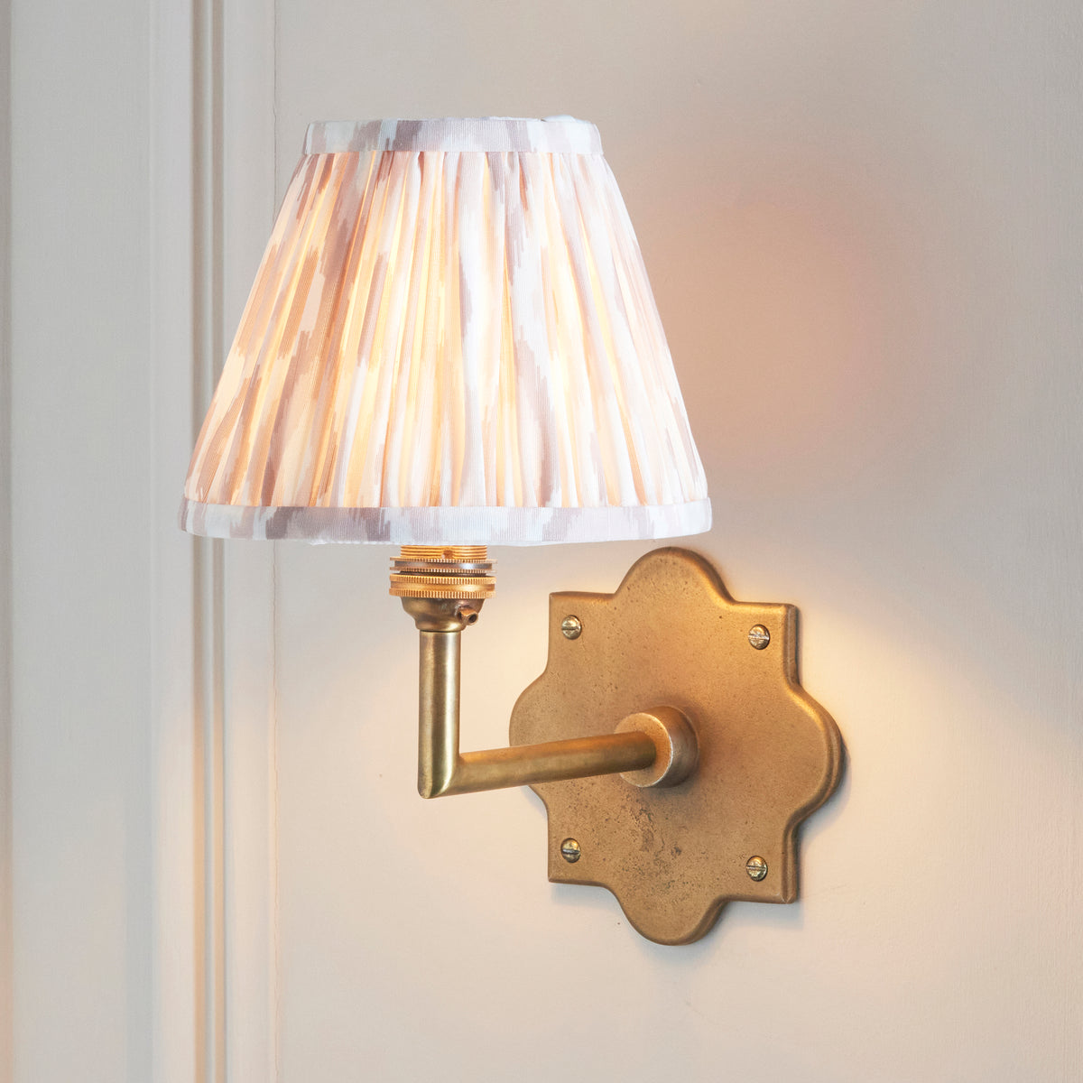 Anvil Angle brass wall light lit on cream panelling, pale ikat pleated shade glowing softly