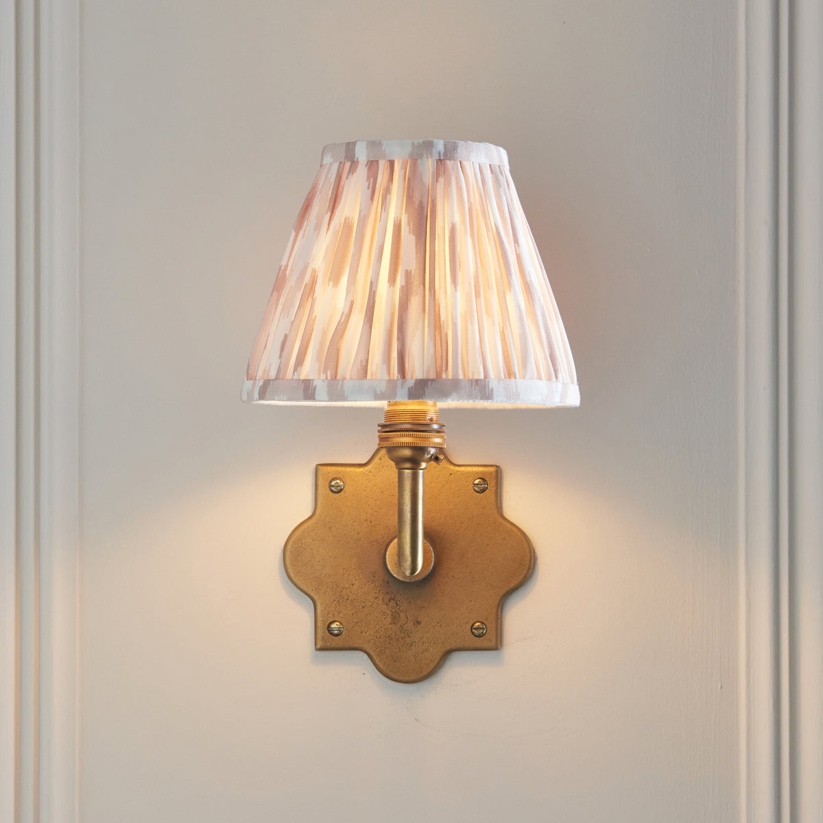 Front view of the Anvil Angle wall light, lit natural ikat shade on a quatrefoil brass backplate