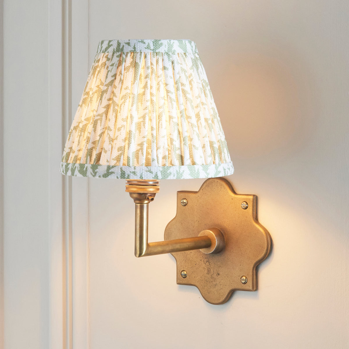 Anvil Angle & Leaf brass wall light lit on cream panelling, green foliage pleated shade glowing