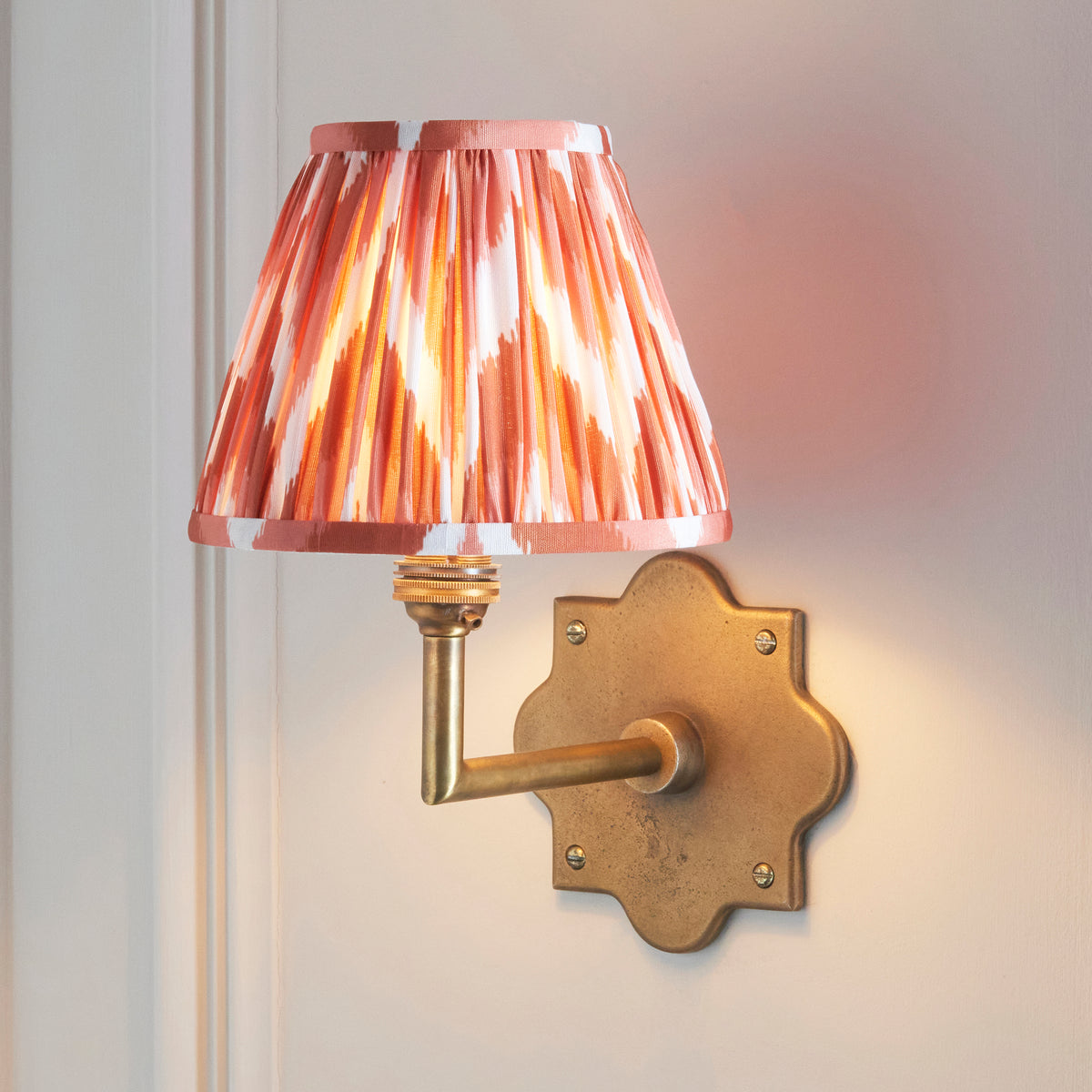 Anvil Angle brass wall light lit on cream panelling, coral zigzag ikat pleated shade glowing