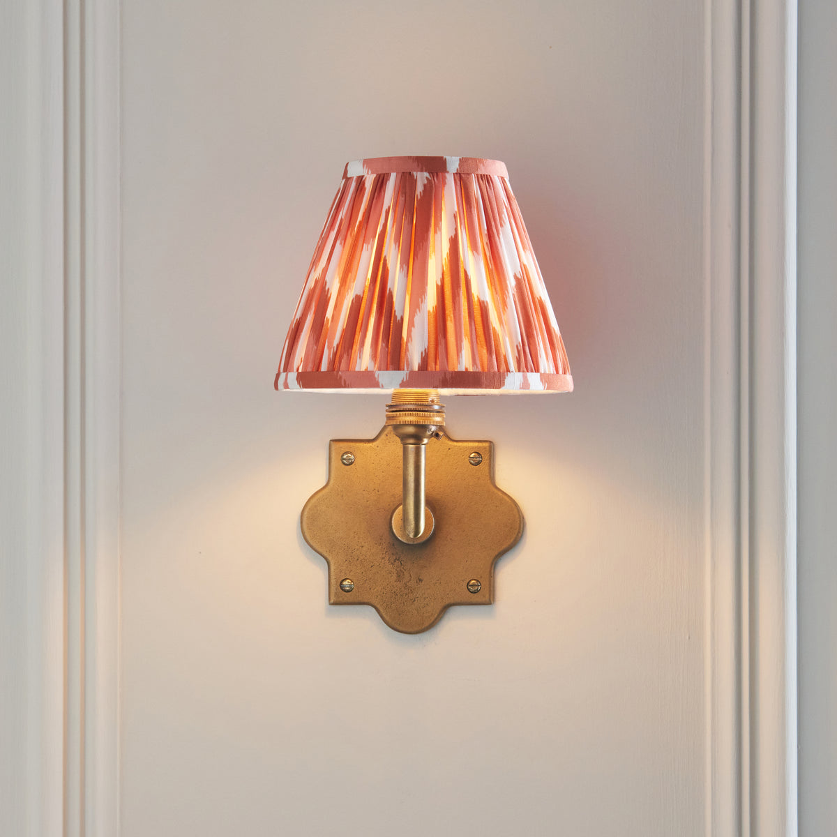 Front view of the Anvil Angle wall light, lit orange zigzag ikat shade on a quatrefoil brass plate
