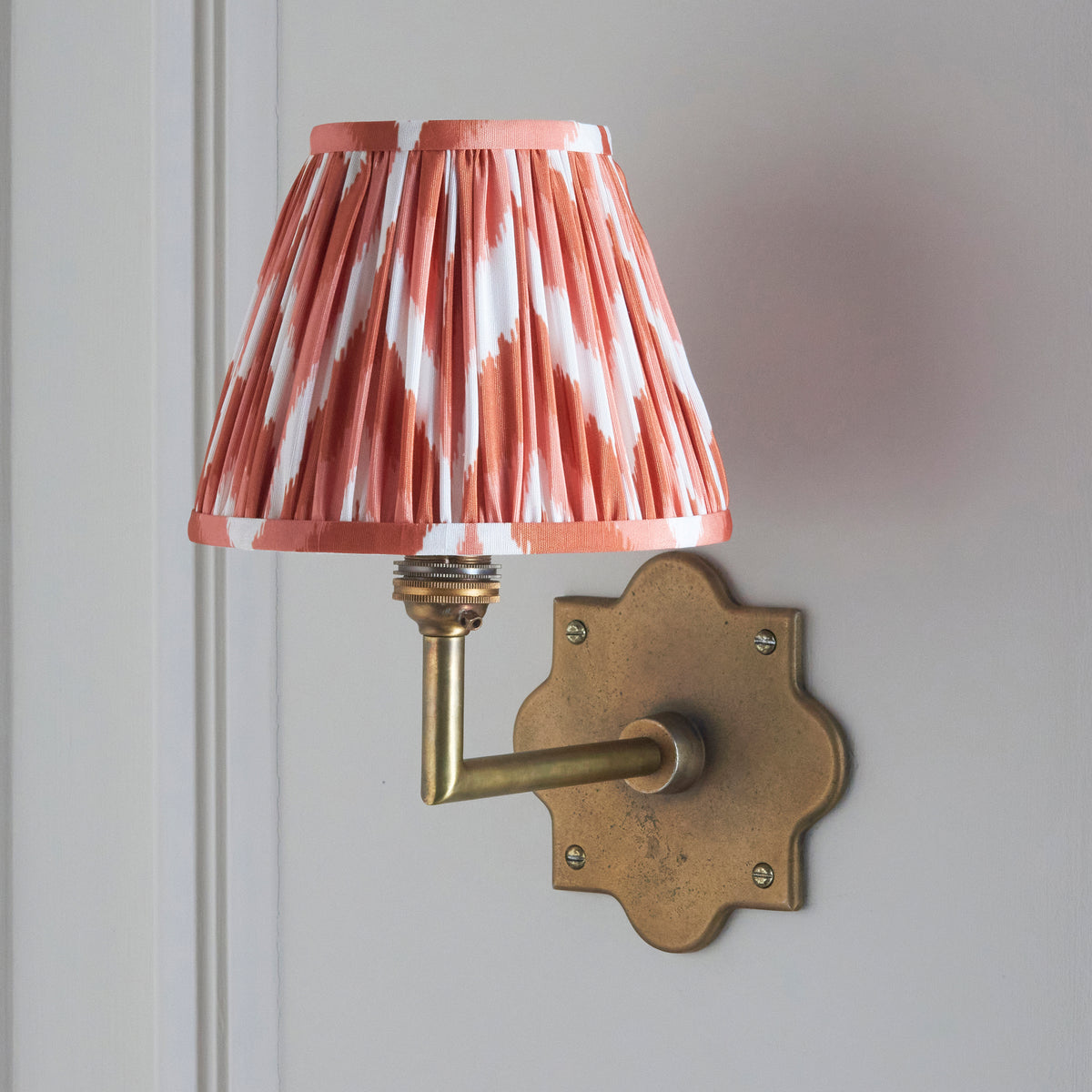 Anvil Angle wall light on pale panelling, coral zigzag ikat pleated shade above an aged brass arm