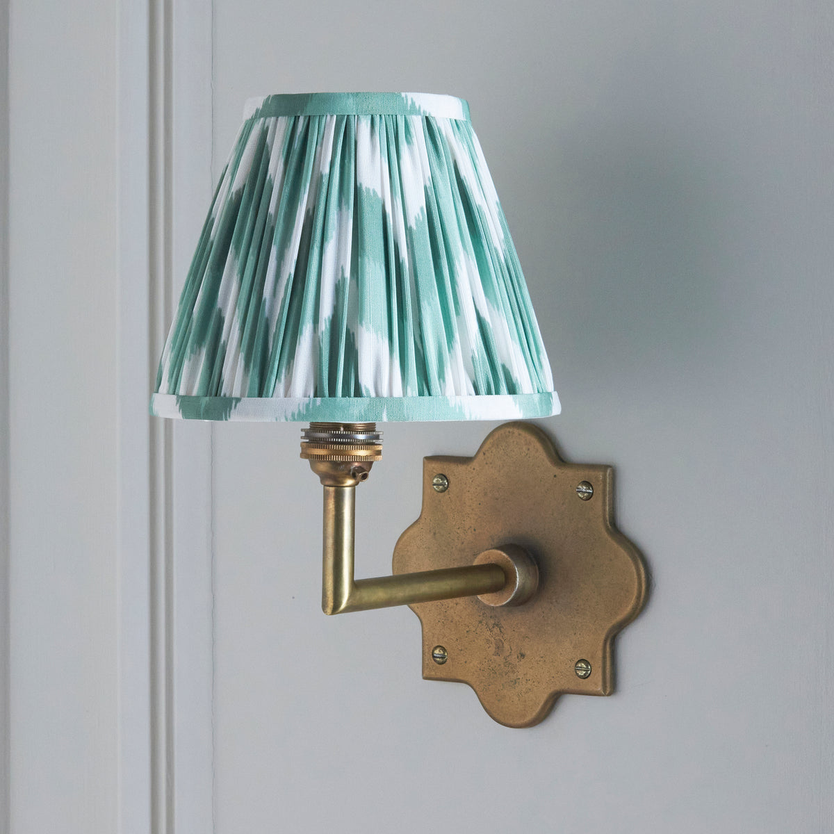 Anvil Angle brass wall light on a grey wall, softer green and white ikat pleated shade illuminated