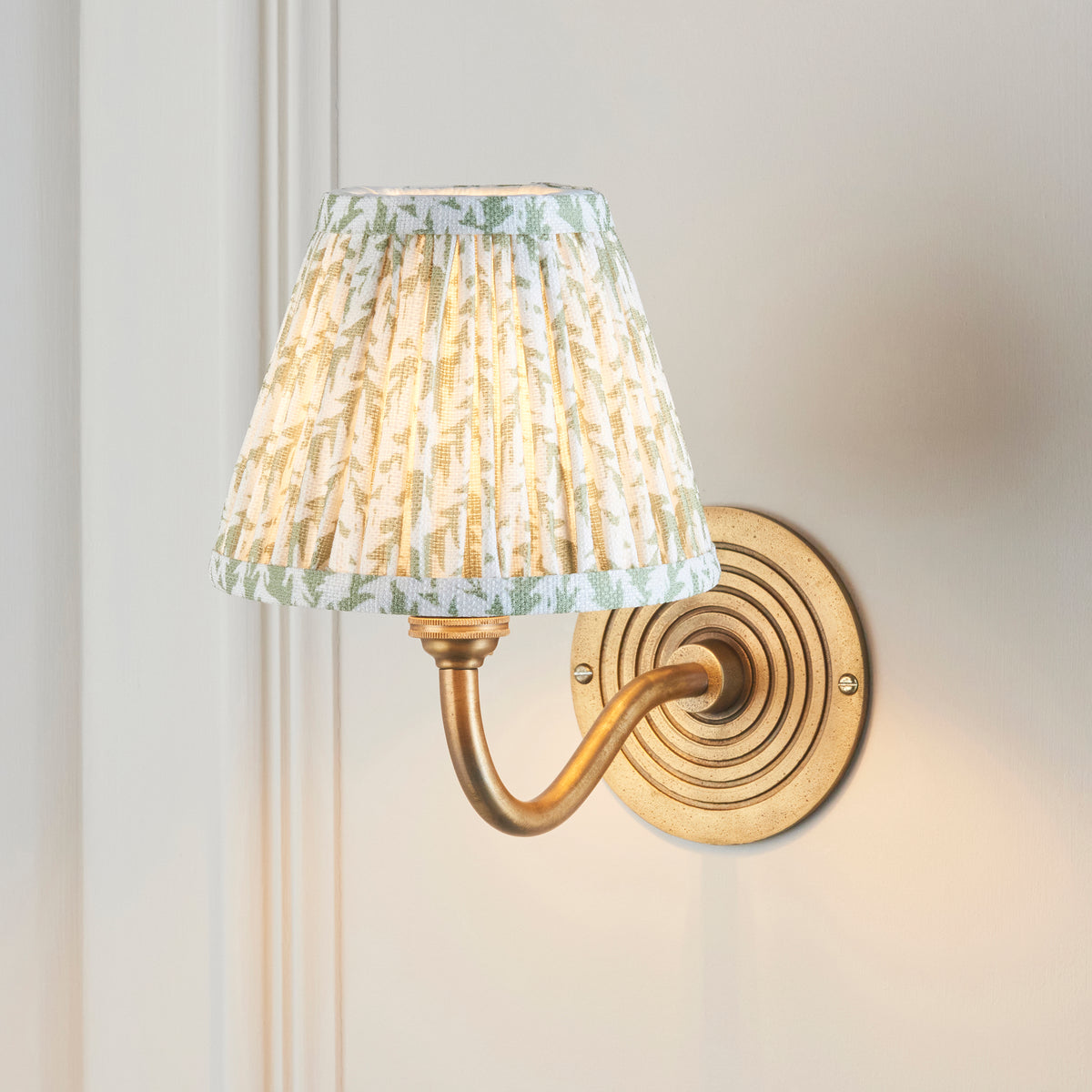 Ripple Curve & Leaf brass wall light lit on panelled off-white wall, leaf-print pleated shade glowing
