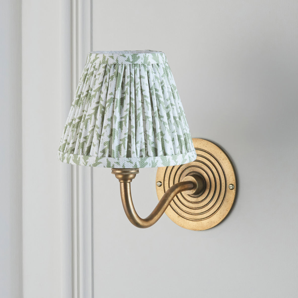 Ripple Curve & Leaf brass wall light beside white panelling, pleated green leaf shade lit softly