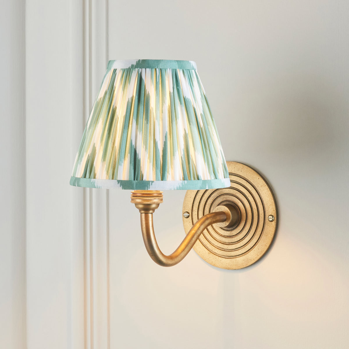 Ripple Curve brass wall light lit on panelled wall, green and cream zigzag ikat pleated shade