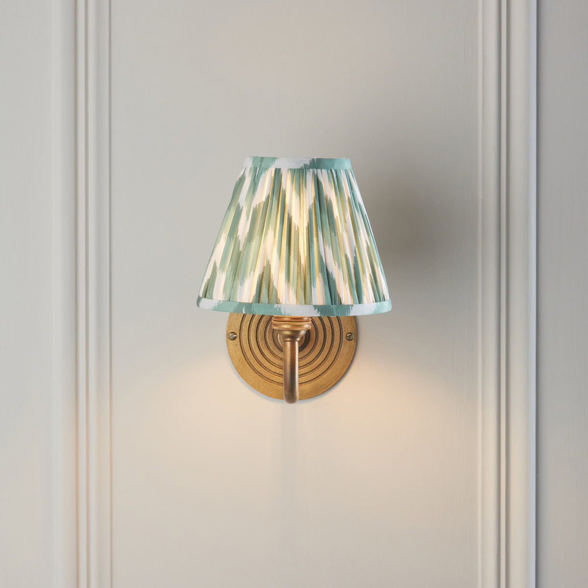 Ripple Curve brass wall light head-on against pale panelling, glowing zigzag ikat shade