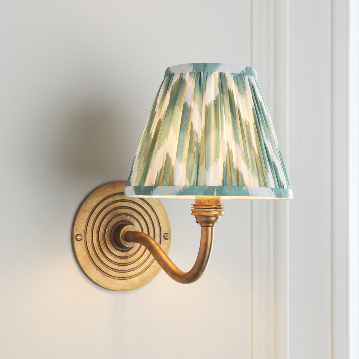 Ripple Curve antique brass wall light angled on off-white wall with pleated green ikat shade