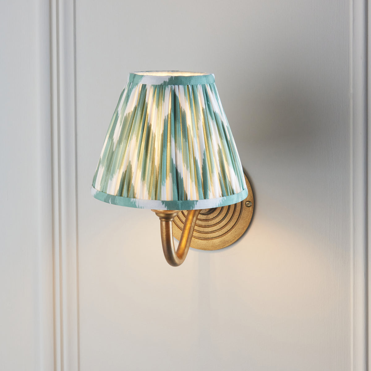 Ripple Curve brass wall light lit on a pale wall, pleated green zigzag ikat shade viewed from below